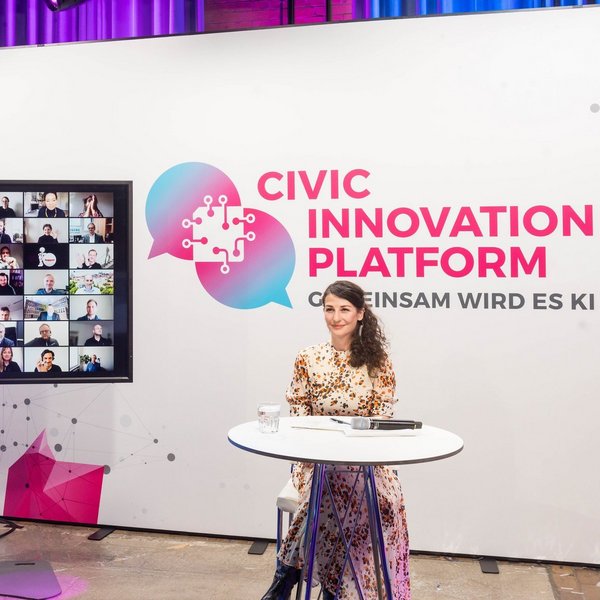 A woman sits at a small round table with a tablet in front of her. Behind her is a large sign that reads Civic Innovation Platform. A screen shows multiple people in a virtual meeting.
