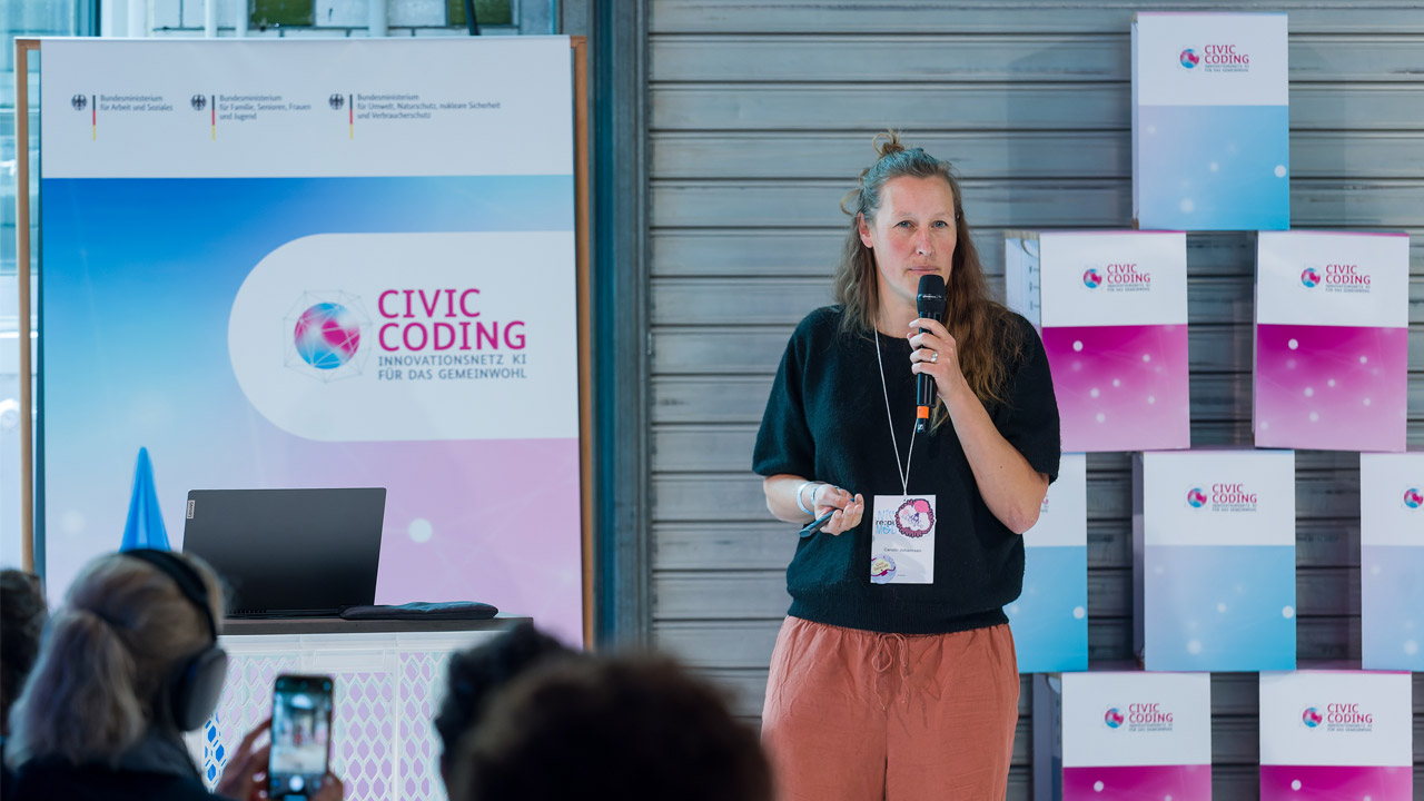 Auf der Bühne ist im Hintergrund links ein Banner von Civic Coding zu sehen, daneben gestapelte Würfel mit dem Logo und im Farbverlauf von Civic Coding. Eine der Gründerinnen steht mit einem Mikrofon darauf, während unten im Bild die Köpfe des Publikums zu erkennen sind.
