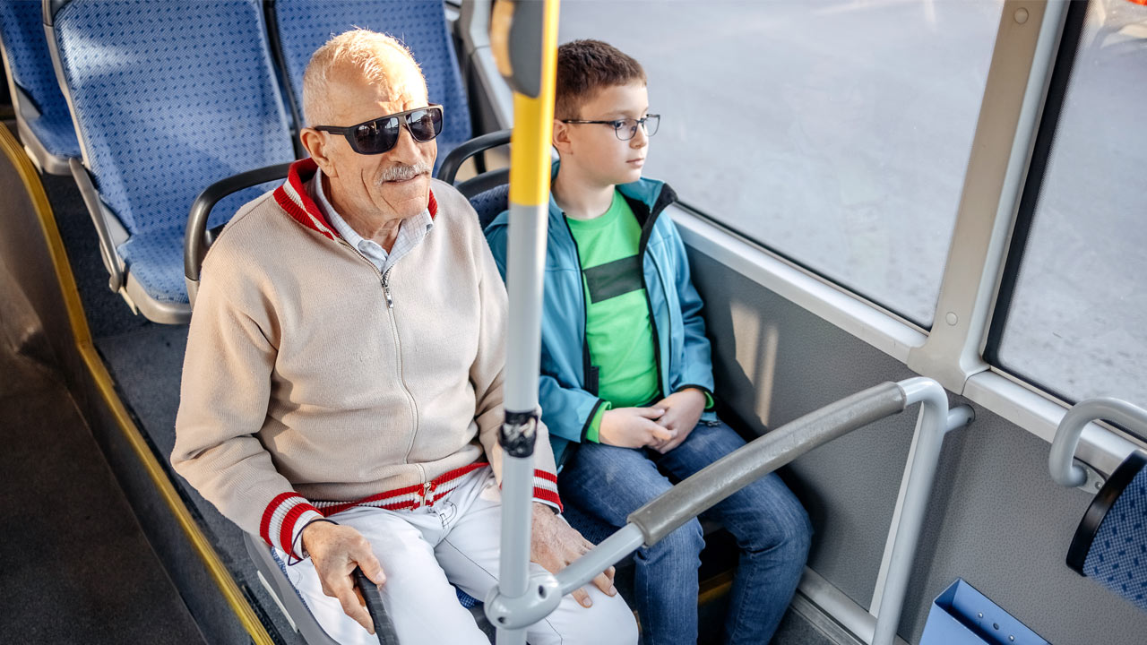 Ein Mann mit Sonnenbrille und Blindenstock sitzt neben einem Kind im Bus. Der Mann lächelt, während das Kind aus dem Fenster schaut.